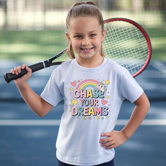 Girl with a tennis racket wearing an adorable girls' tennis t-shirt featuring a cute unicorn, colorful rainbow, and "Chase Your Dreams" slogan with tennis ball accents.