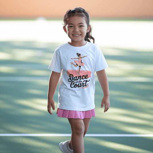 Young girl wearing a stylish girls' tennis t-shirt featuring a graceful young ballerina in a pink tutu performing an elegant pose on a tennis court. The text 'Dance on the Court' blends ballet and tennis, celebrating the fluid movement and grace of the game.
