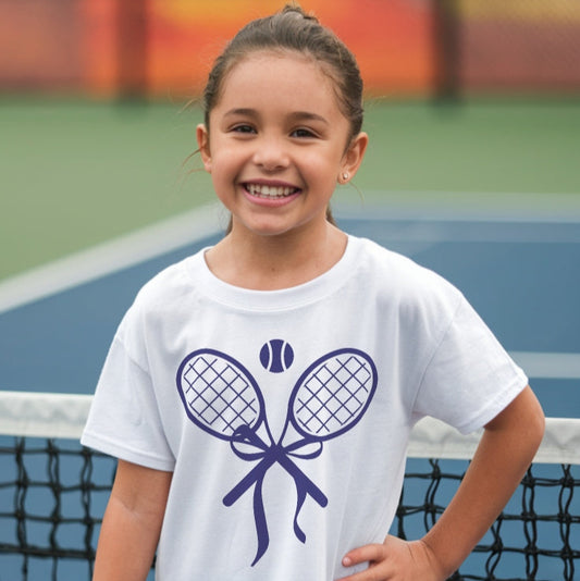 Young girl wearing a cute girls' tennis t-shirt in white featuring a stylish design of two crossed tennis rackets tied with a bow and a tennis ball above.
