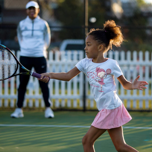 Girl performing a tennis stroke and wearing a white tennis t-shirt featuring a charming illustration of a ballerina holding a tennis racket, with the words "Grace, Grit, and Game" in pink.