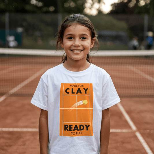 Young girl standing on the clay tennis court, wearing a white t-shirt featuring a design of a tennis court and a slogan "Made for Clay, Ready to Play"