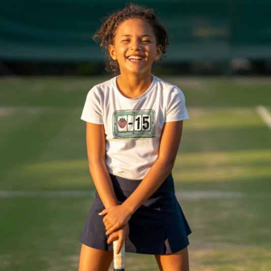 Young girl standing on a grass tennis court and wearing a white t-shirt with strawberry tennis scoreboard design showing “-15”