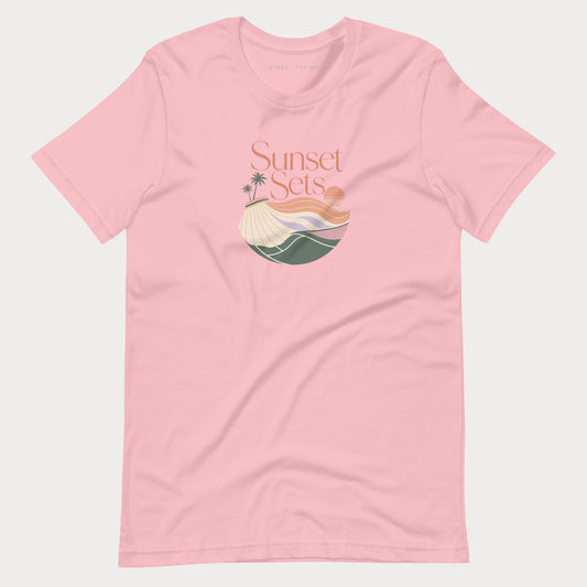 A light pink short-sleeve crewneck women's lifestyle tennis t-shirt featuring a retro-style graphic on the chest with the text 'Sunset Sets' above a stylized beach scene with palm trees, a sun, and waves.