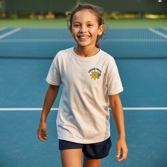 Young girl on a tennis court, wearing girls tennis t-shirt with smiling sunshine character and Sunshine Smash slogan.