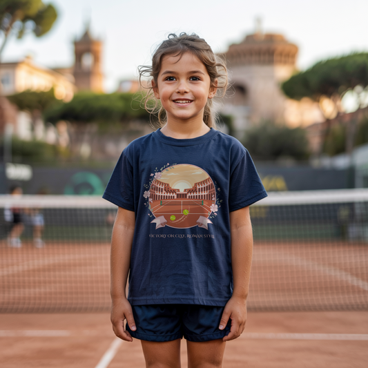 Young girl standing on a tennis court and wearing a navy T-shirt featuring a clay court set inside an ancient Roman amphitheater, surrounded by floral accents and a banner reading "Victory on Clay, Roman Style."