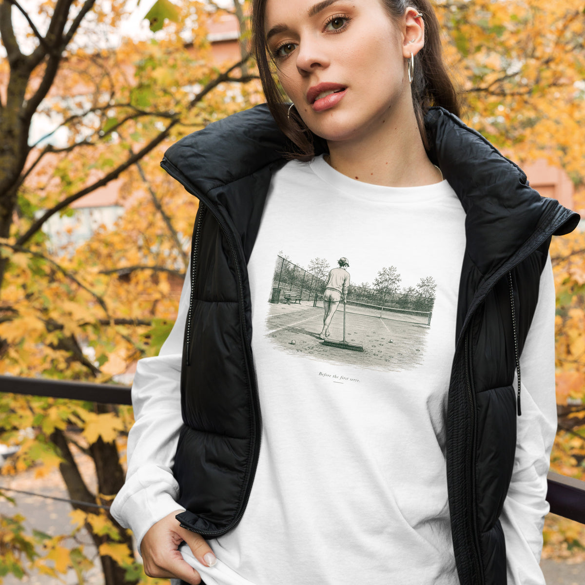 Woman in a black puffer vest among autumn foliage wearing a white Before the First Serve long-sleeve T-shirt with a pen-and-ink tennis court illustration