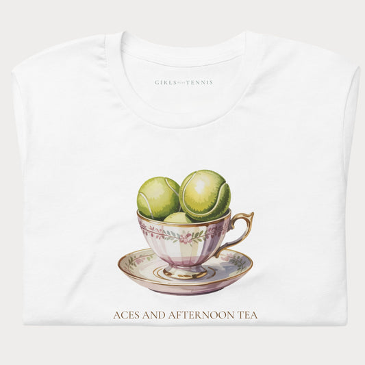 A folded white t-shirt with a vintage-style graphic of green tennis balls in a pink teacup on a saucer, with the text 'GIRLS-TENNIS' inside the collar and 'ACES AND AFTERNOON TEA' printed below.
