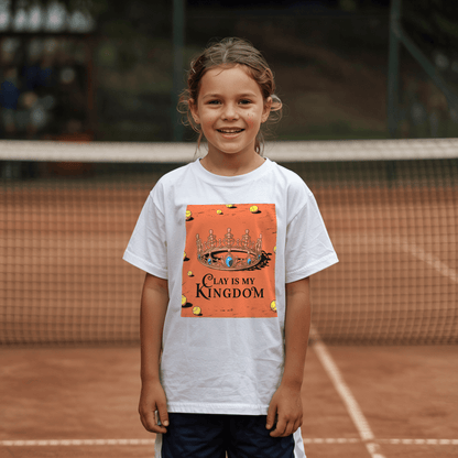 Young girl standing on a clay tennis court and wearing a white tennis T-shirt featuring a regal crown on a clay court with scattered tennis balls and the phrase "Clay Is My Kingdom" in bold lettering
