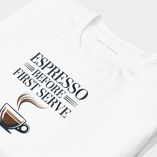 A close-up of a white short-sleeve tennis lifestyle t-shirt featuring a graphic print that reads 'ESPRESSO BEFORE FIRST SERVE' in dark lettering, accompanied by an illustration of an espresso cup with steam. The inside collar shows the brand name 'GIRLS-PLAY-TENNIS'.