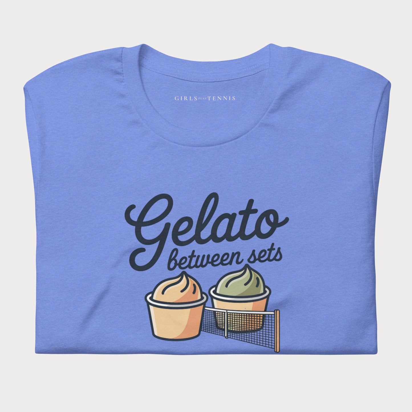 A folded blue t-shirt with a graphic print that reads 'Gelato between sets' in black script, accompanied by two illustrated cupcakes and a tennis net. The inner collar shows the brand name 'GIRLS PLAY TENNIS'.