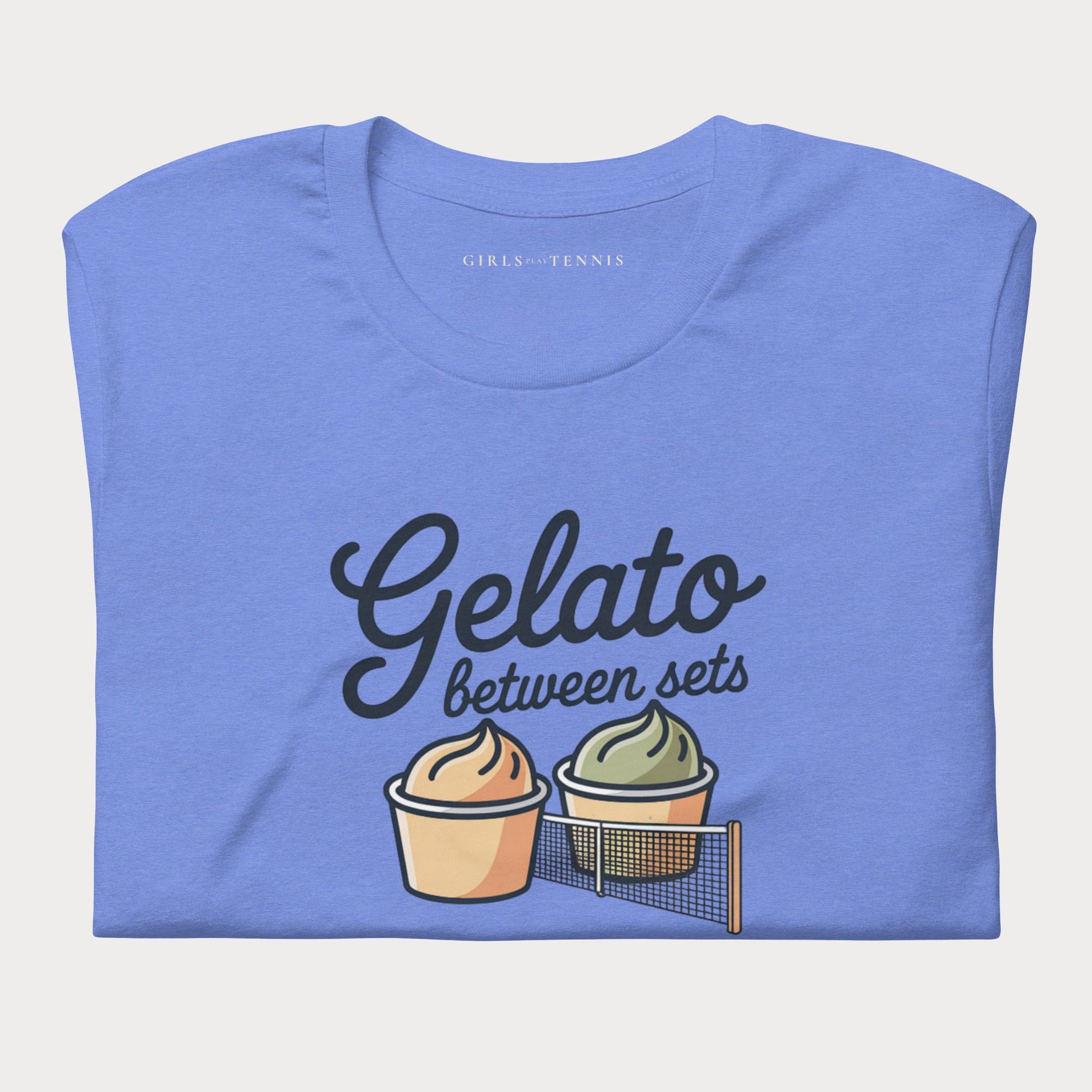 A folded blue t-shirt with a graphic print that reads 'Gelato between sets' in black script, accompanied by two illustrated cupcakes and a tennis net. The inner collar shows the brand name 'GIRLS PLAY TENNIS'.