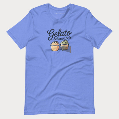 A heather blue short-sleeve women's tennis t-shirt with a graphic on the chest that reads 'Gelato between sets' above two illustrated gelato cups, shown on a light gray background.