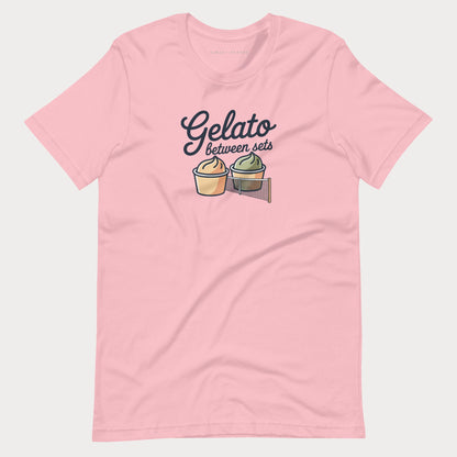 A light pink short-sleeve women's tennis t-shirt with a graphic on the chest that reads 'Gelato between sets' above two illustrated gelato cones, shown on a light gray background.