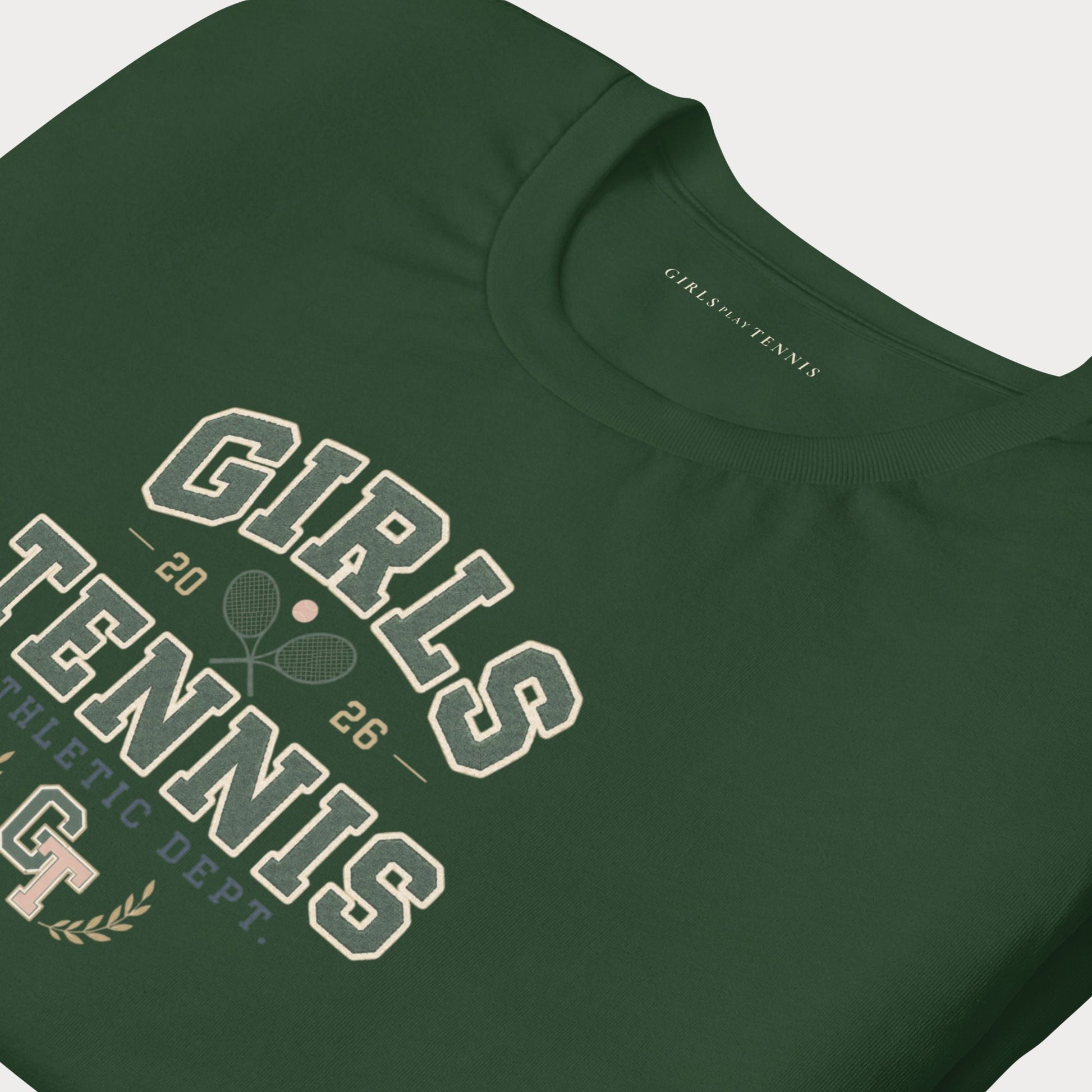 A close-up of a dark green short-sleeve t-shirt featuring a vintage-style collegiate graphic that reads 'GIRLS TENNIS' in cream and white, with crossed tennis rackets, the number '26', and 'ATHLETIC DEPT.' printed on the front.
