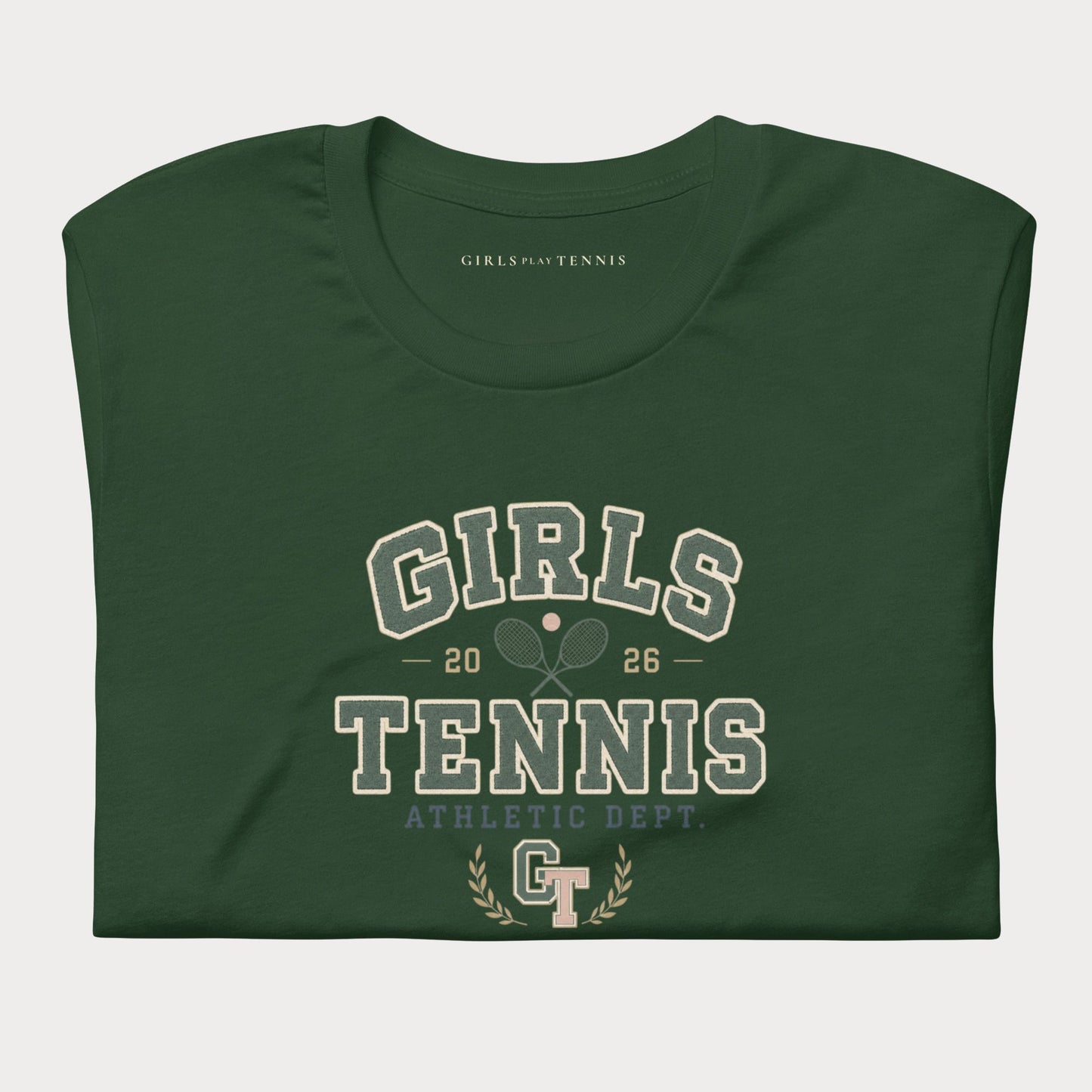 A dark green folded t-shirt with a vintage-style collegiate graphic reading 'GIRLS TENNIS' and 'ATHLETIC DEPT.' in cream and light blue-gray lettering, on a light gray background.