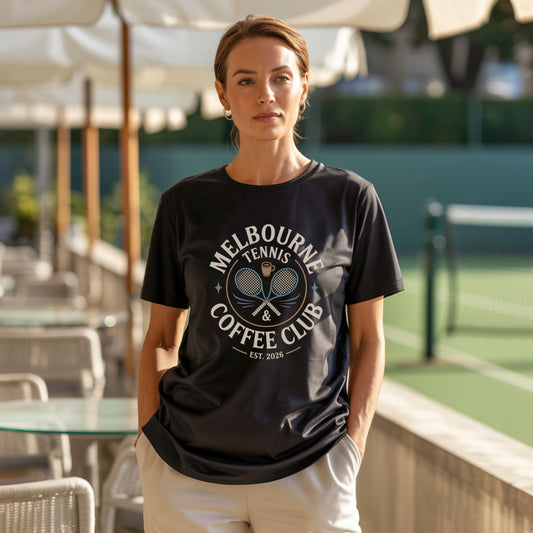 A women standing in a coffee shop by a tennis court, wearing a Melbourne Tennis & Coffee Club t-shirt with crossed racquets and coffee cup for adults and older youth.