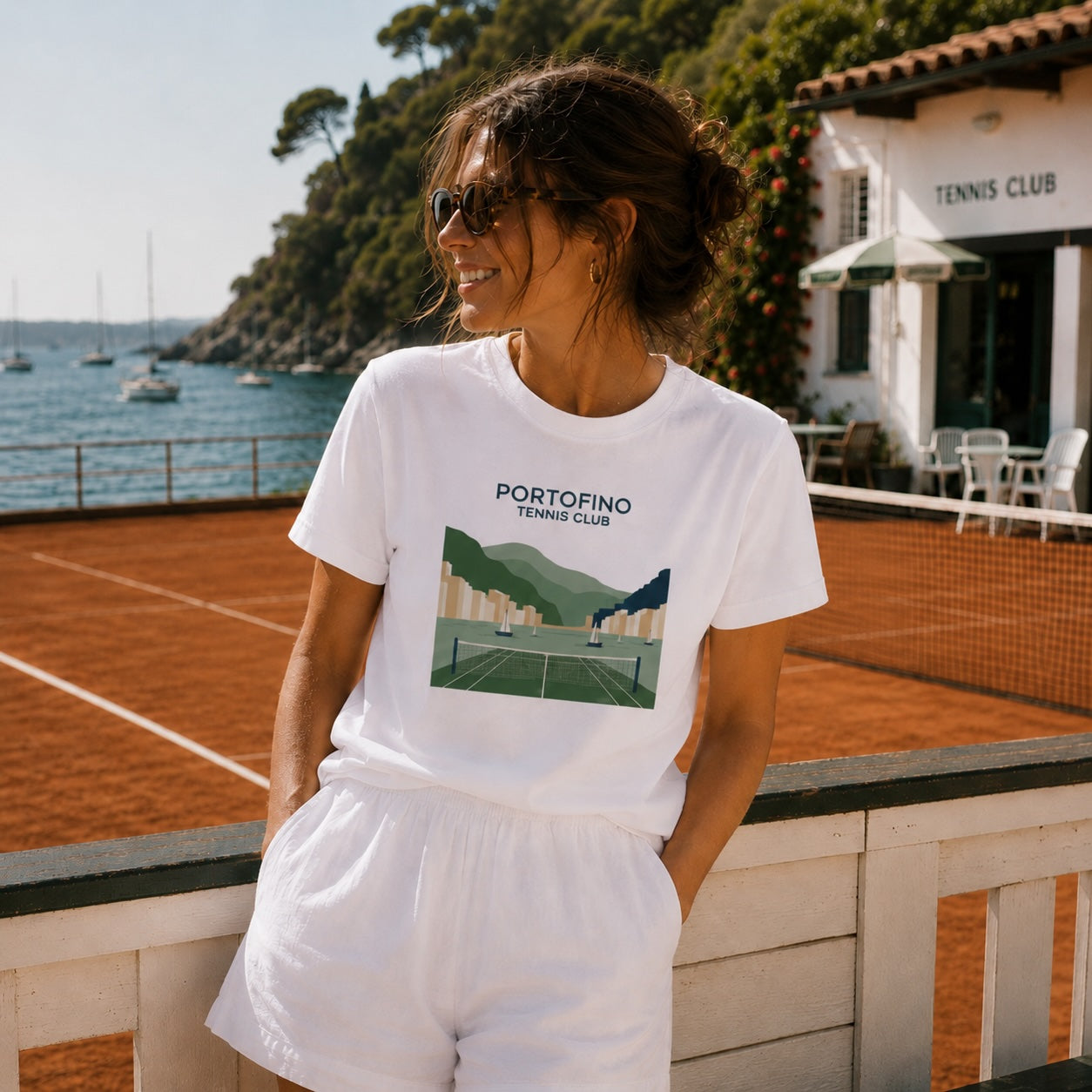A woman wearing a white graphic t-shirt and white shorts stands on a red clay tennis court with a sea and building in the background. The t-shirt features a green and blue tennis court graphic with the text 'PORTOFINO TENNIS CLUB'.