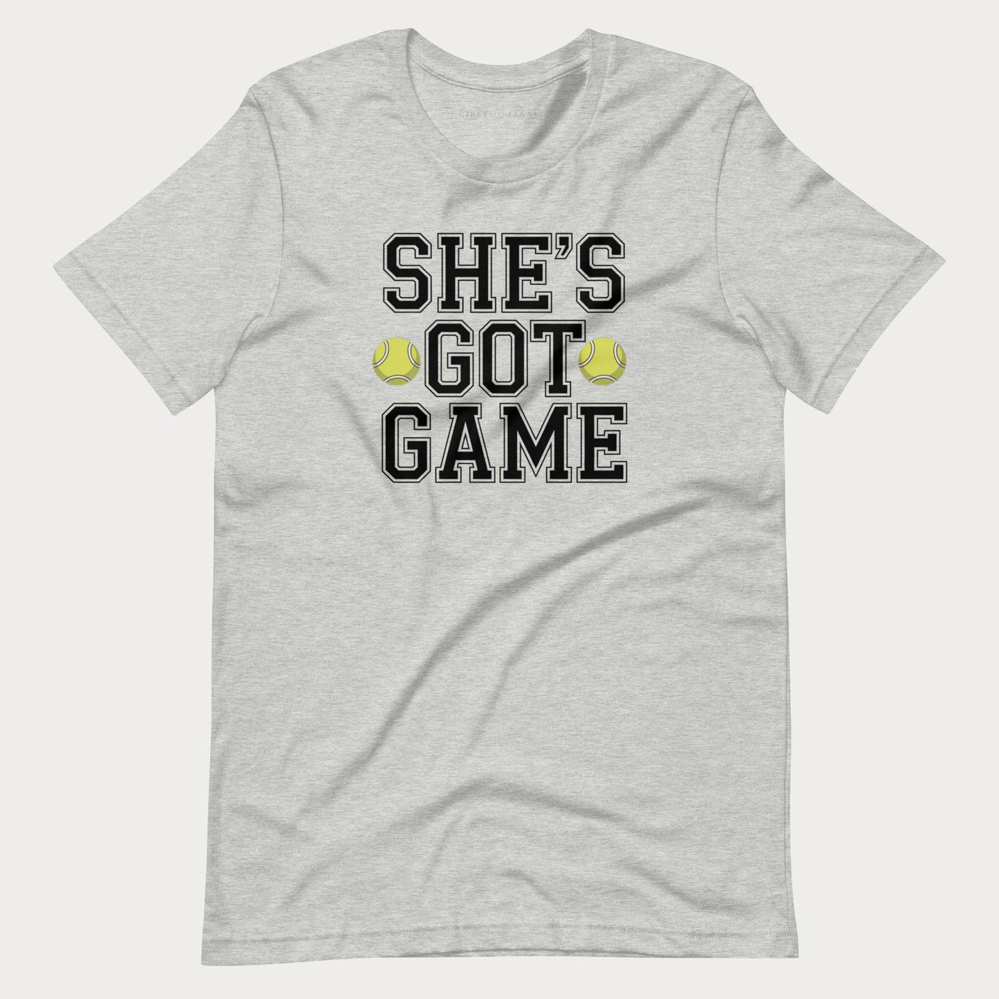 A light heather gray short-sleeve women's tennis t-shirt with a graphic print on the front that reads 'SHE'S GOT GAME' in black varsity-style letters, accented by two yellow tennis balls.
