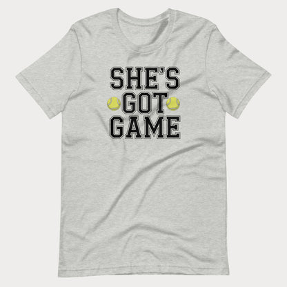 A light heather gray short-sleeve women's tennis t-shirt with a graphic print on the front that reads 'SHE'S GOT GAME' in black varsity-style letters, accented by two yellow tennis balls.