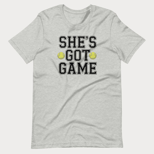 A light heather gray short-sleeve women's tennis t-shirt with a graphic print on the front that reads 'SHE'S GOT GAME' in black varsity-style letters, accented by two yellow tennis balls.