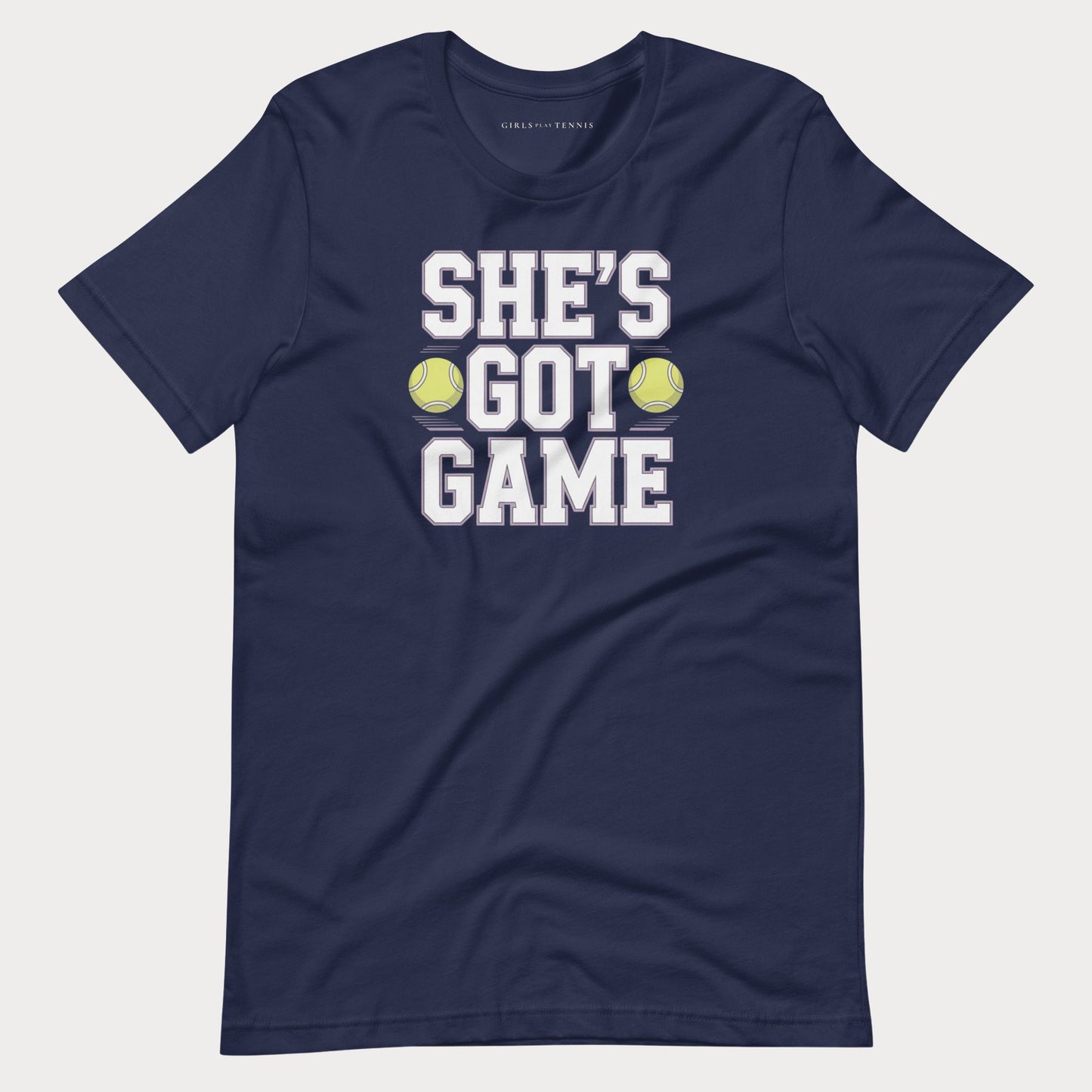 A navy blue short-sleeve women's tennis t-shirt with a graphic print that reads 'SHE'S GOT GAME' in white block letters, accented by two yellow tennis balls, shown on a light gray background.