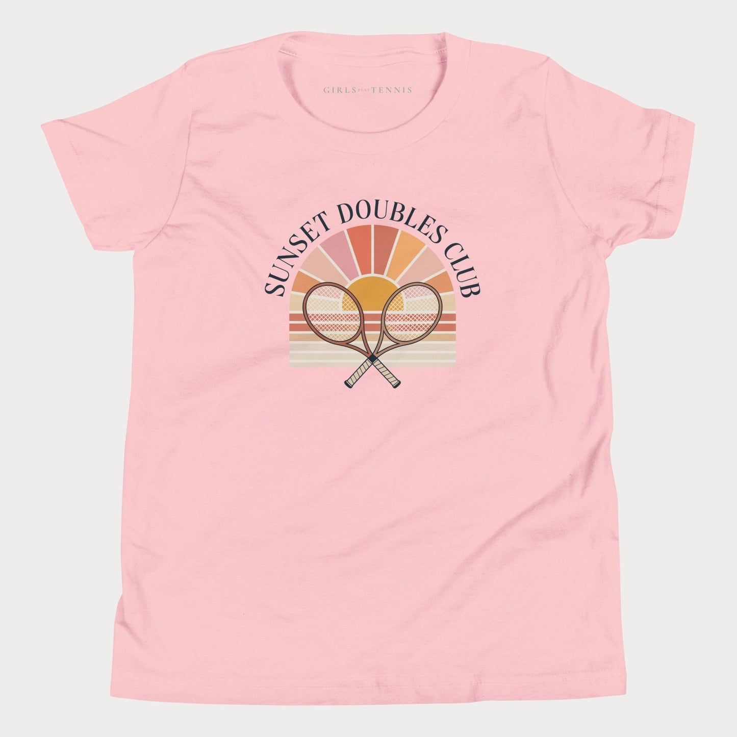 A light pink short-sleeve girls tennis t-shirt with a retro-style graphic on the front. The graphic features the text 'SUNSET DOUBLES CLUB' arched over a stylized sunburst with two crossed tennis rackets