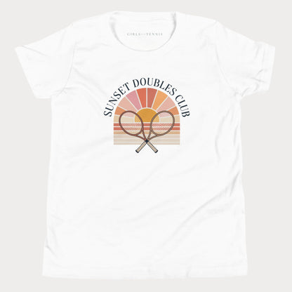 A white short-sleeve crewneck girls tennis t-shirt featuring a retro-style graphic on the front with two crossed tennis rackets in front of a sunset gradient and the text 'SUNSET DOUBLES CLUB'. The inner neck label reads 'GIRLS PLAY TENNIS'.
