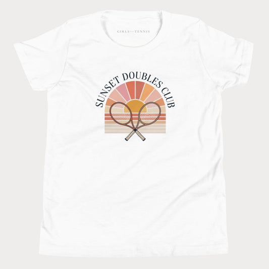 A white short-sleeve crewneck girls tennis t-shirt featuring a retro-style graphic on the front with two crossed tennis rackets in front of a sunset gradient and the text 'SUNSET DOUBLES CLUB'. The inner neck label reads 'GIRLS PLAY TENNIS'.
