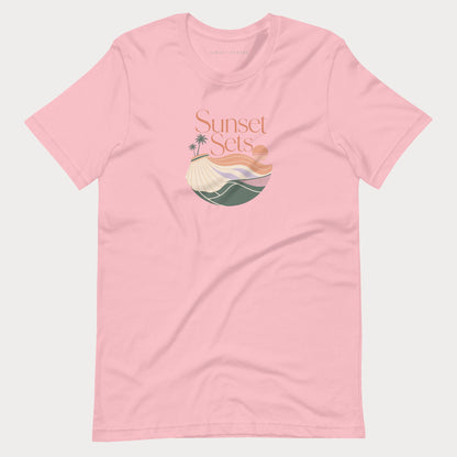 A light pink short-sleeve crewneck women's lifestyle tennis t-shirt featuring a retro-style graphic on the chest with the text 'Sunset Sets' above a stylized beach scene with palm trees, a sun, and waves.