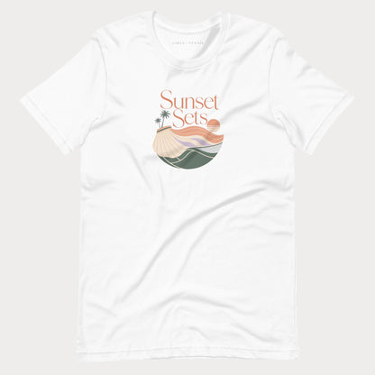 A white short-sleeve crewneck women's lifestyle tennis t-shirt featuring a 'Sunset Sets' graphic on the chest with a beach, palm trees, and waves in muted orange, beige, and green tones, displayed on a light gray background.