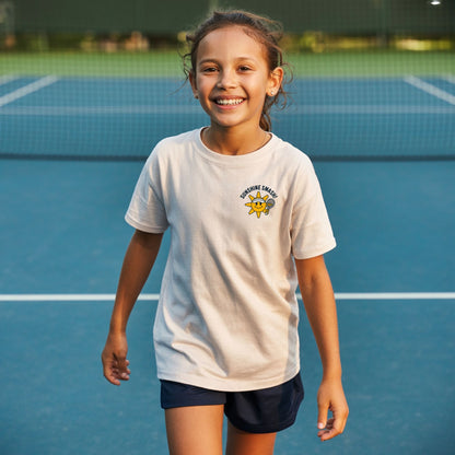 Young girl on a tennis court, wearing girls tennis t-shirt with smiling sunshine character and Sunshine Smash slogan.