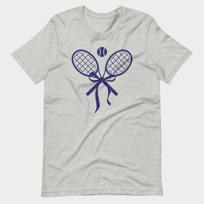 A light heather gray short-sleeve tennis t-shirt with a navy blue graphic of two crossed tennis rackets, a tennis ball, and a ribbon bow on the chest, displayed against a light gray background.