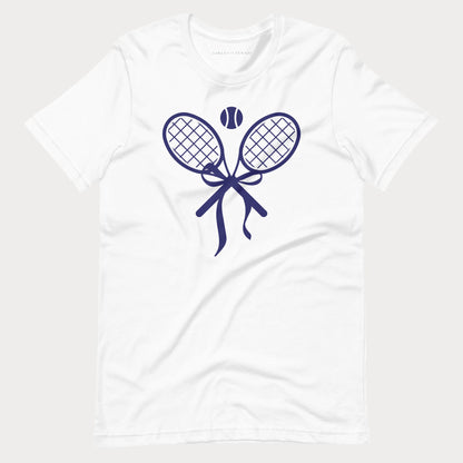 A white short-sleeve crewneck tennis t-shirt featuring a navy blue graphic of two tennis rackets tied in a bow with a tennis ball above them, displayed on a light gray background.