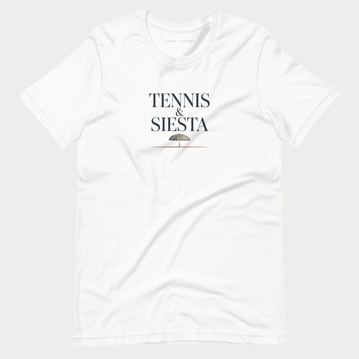 A white short-sleeve crewneck women's lifestyle t-shirt with 'TENNIS & SIESTA' printed in black serif letters on the chest, with a small umbrella graphic underneath, shown against a light gray background.