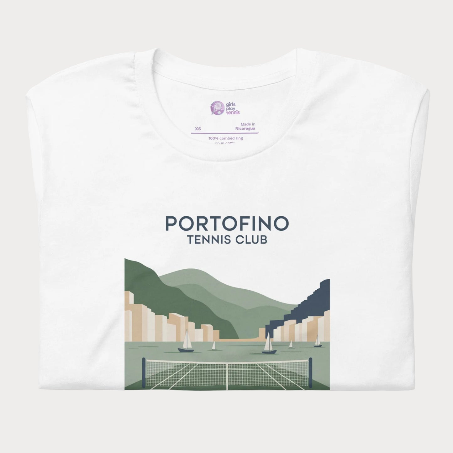 A folded white t-shirt with a graphic print of a tennis club in Portofino, featuring green hills, sailboats, and a tennis court, displayed on a clean white background.