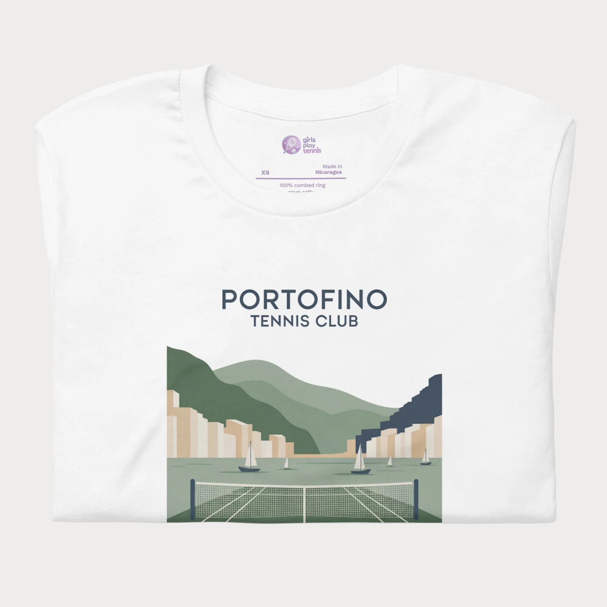 A folded white t-shirt with a graphic print of a tennis club in Portofino, featuring green hills, sailboats, and a tennis court, displayed on a clean white background.