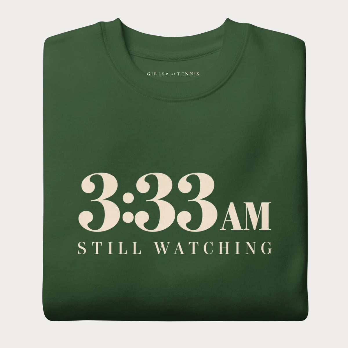 Folded forest green 3:33 AM women's tennis crewneck sweatshirt with cream lettering reading 3:33 AM above Still Watching