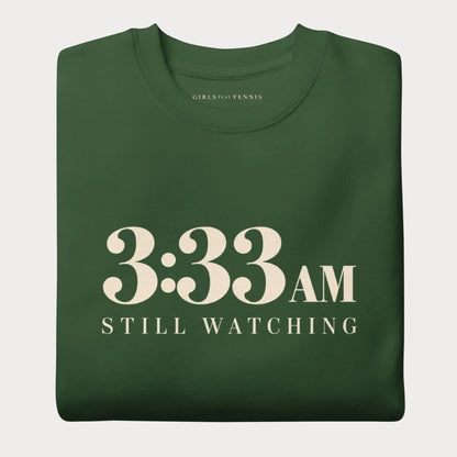 Folded forest green 3:33 AM women's tennis crewneck sweatshirt with cream lettering reading 3:33 AM above Still Watching