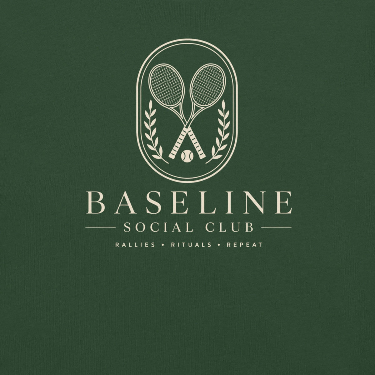 Close-up of the Baseline Social Club crest with crossed rackets, laurel and "Rallies • Rituals • Repeat" on a forest green tee