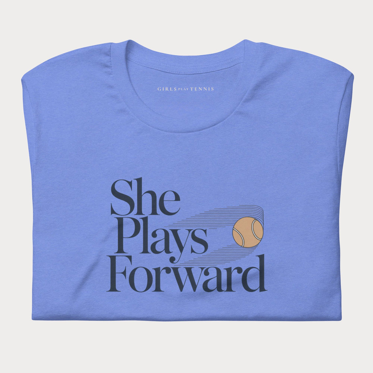 Folded blue She Plays Forward women's tennis T-shirt with stacked lettering and a motion-blurred tennis ball