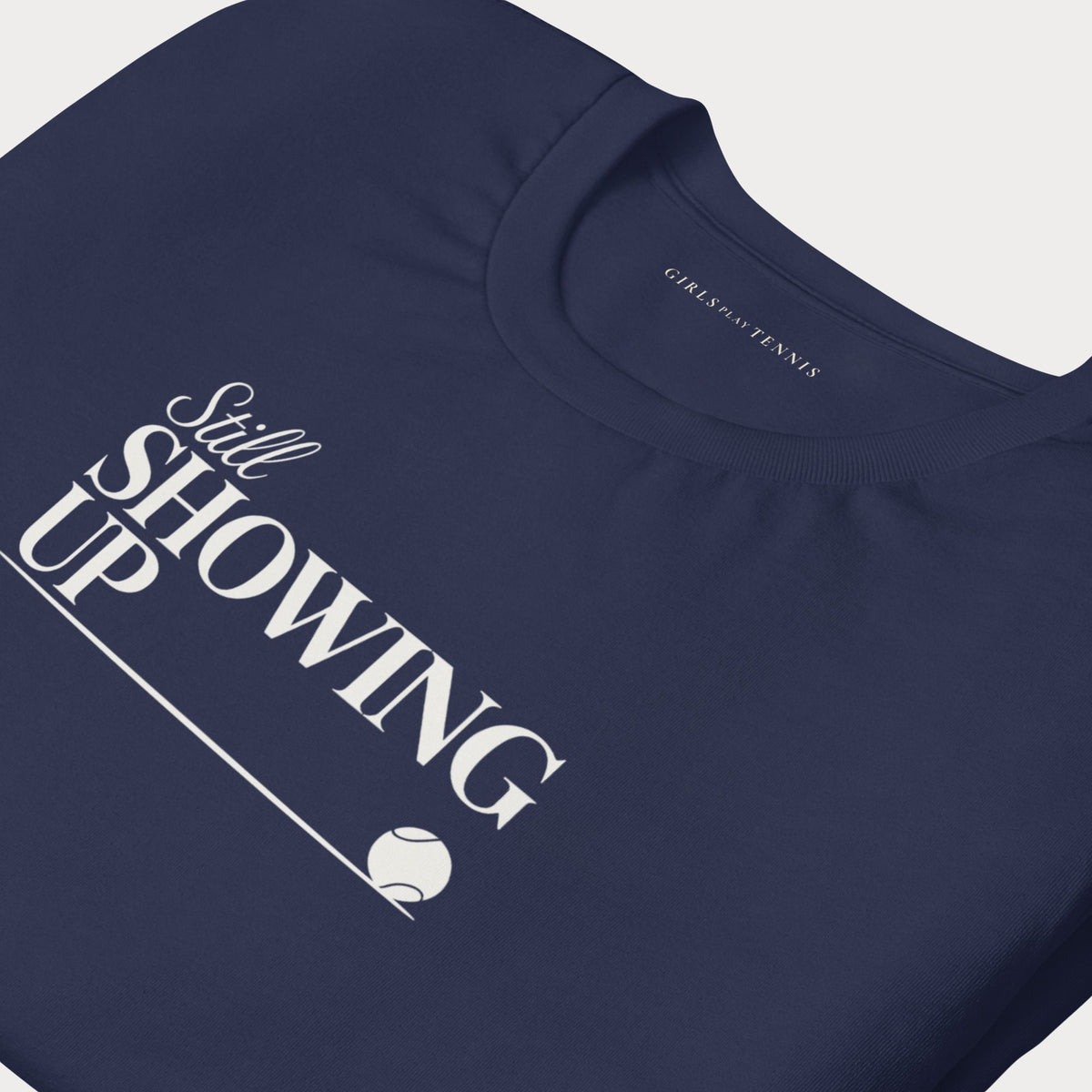 Close-up of the Still Showing Up lettering and tennis ball on a navy tee