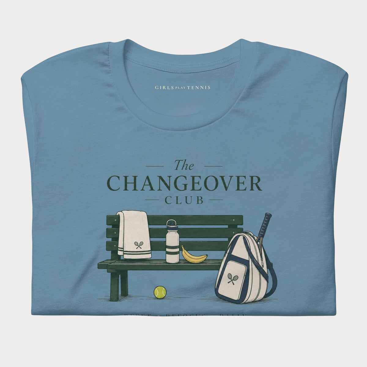 Folded steel blue The Changeover Club women's tennis T-shirt with a courtside bench, towel, racket bag and banana illustration